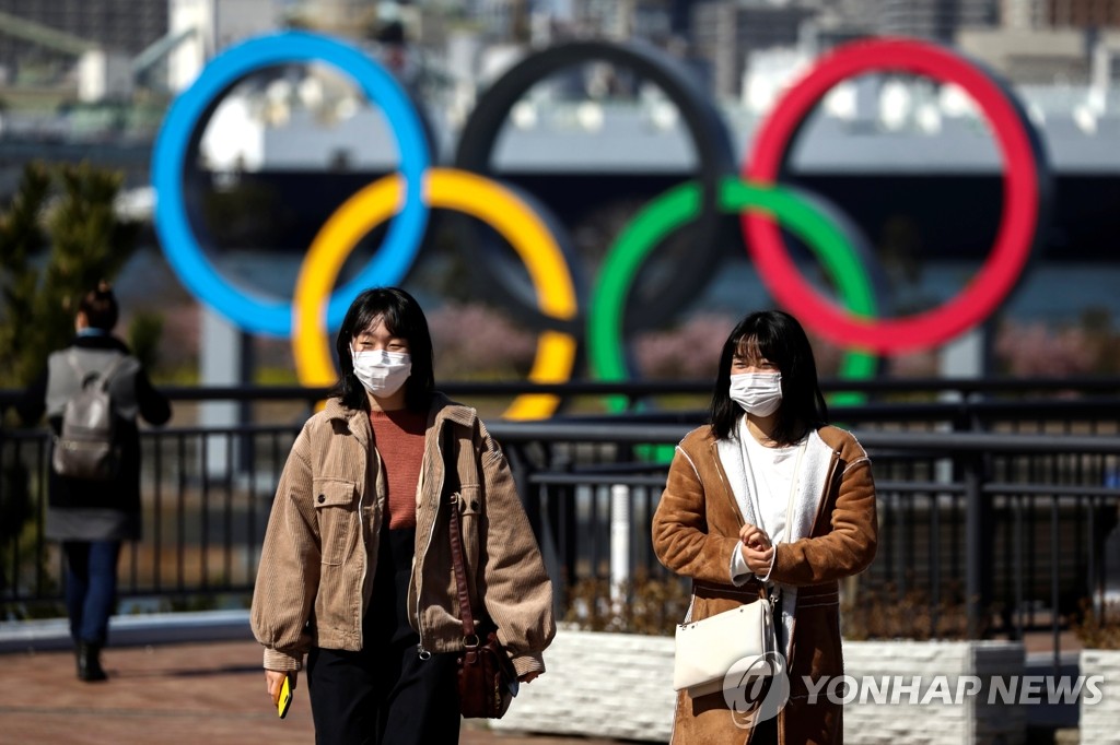 Japanese citizens wearing masks walk outside on Feb. 27, 2020, in front of the Olympic rings installed at the Odaiba Marine Park in Tokyo, ahead of the Tokyo 2020 Summer Olympic Games. (Yonhap)